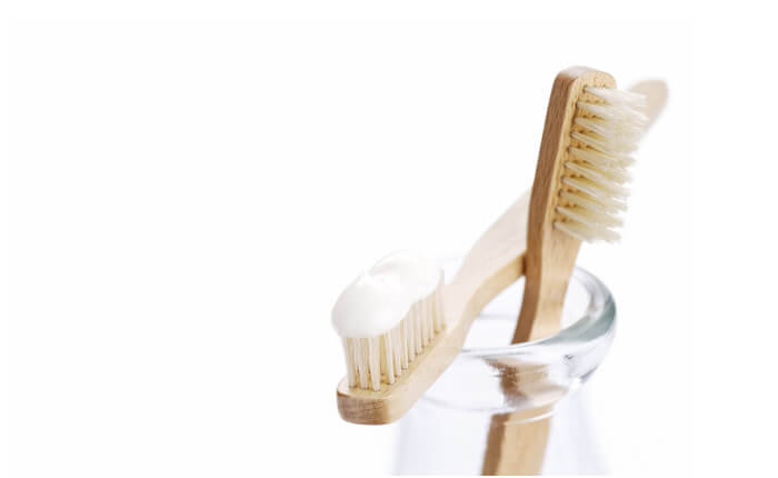 Set of two wooden toothbrushes in a glass jar, one with white toothpaste on it, in front of a white background.