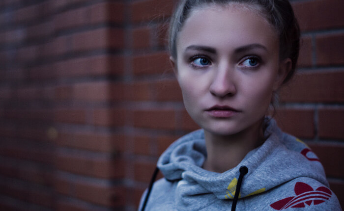 Blonde young woman wearing a gray sweatshirt looks anxiously away as she stands against a red brick wall