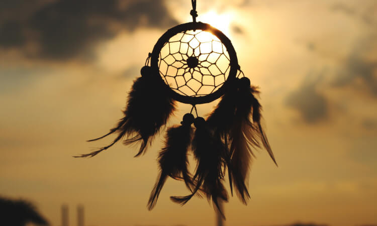 Silhouette of a woven dreamcatcher with feathers against a golden sky