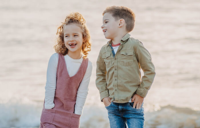 A blonde girl wearing a pink dress stands next to a blonde boy in jeans, both with their hands in their pockets